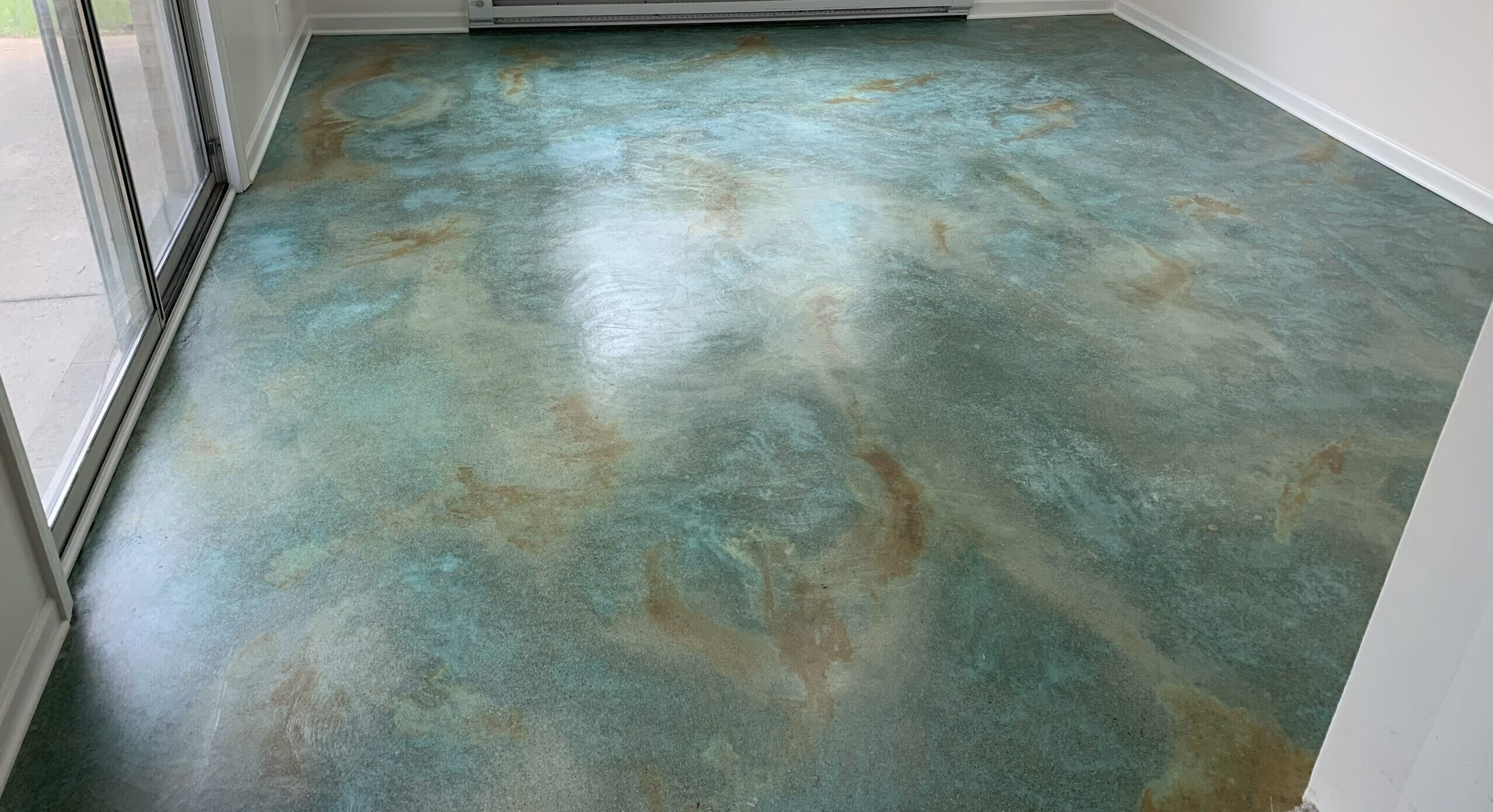 A room with a polished concrete floor featuring a marbled blend of blue and earthy brown tones created with an acid stain. Sunlight from large sliding glass doors highlights the sheen from a satin sealer applied to the floor.