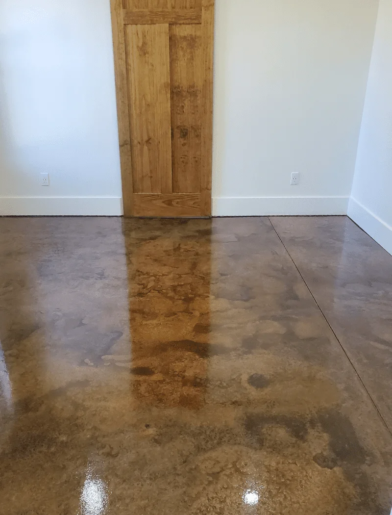 A well-lit room with a glossy, acid-stained concrete floor in varying shades of brown, creating a smooth, marble-like finish that complements the rustic wooden door.
