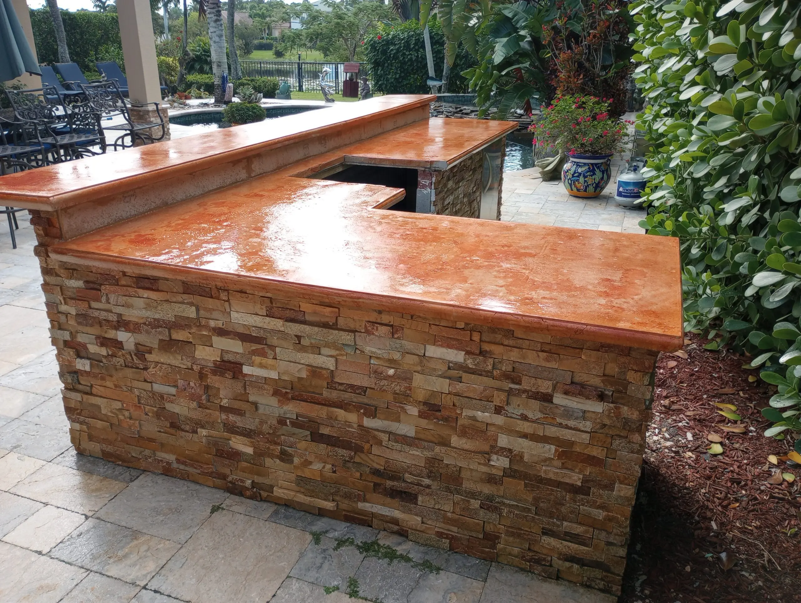 An outdoor countertop stained in ColorWave Sunset, with a stacked stone base and a glossy finish, situated near a pool and garden area.