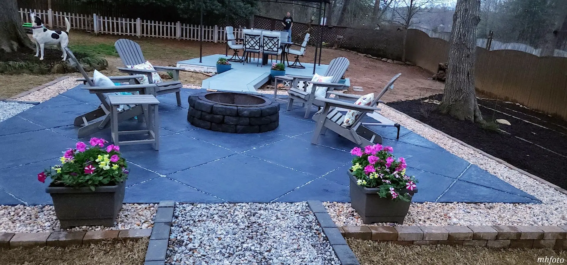 Stamped concrete patio stained in Iron Gray with Adirondack chairs around a fire pit, surrounded by flower pots and gravel landscaping, with a dog in the background.