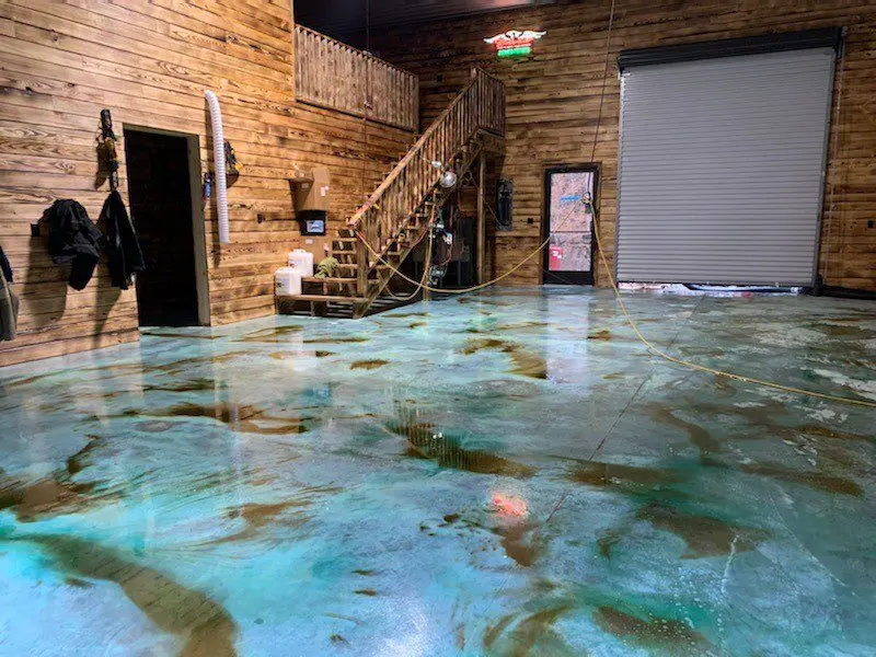 A large garage with a polished concrete floor, showcasing a vibrant blend of azure blue and sandy brown tones. The floor has a marbled, veining effect enhanced by an acid staining technique and the use of Miracle Gro for intensified blue hues.