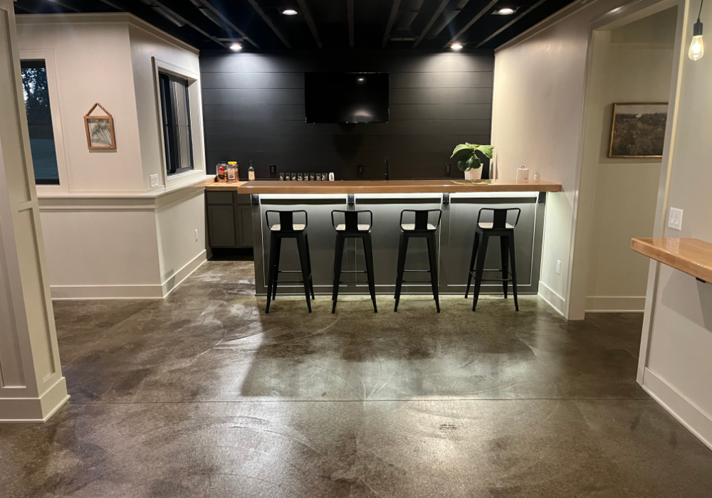 A stylish basement bar area with a sleek stained concrete floor in ColorWave Molasses, featuring industrial-style bar stools