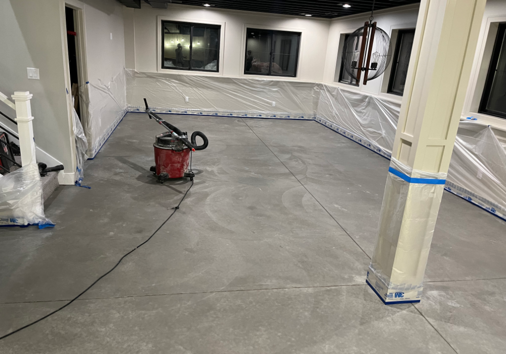 A fully prepped concrete basement floor ready for staining, with protective plastic covering the walls and trim