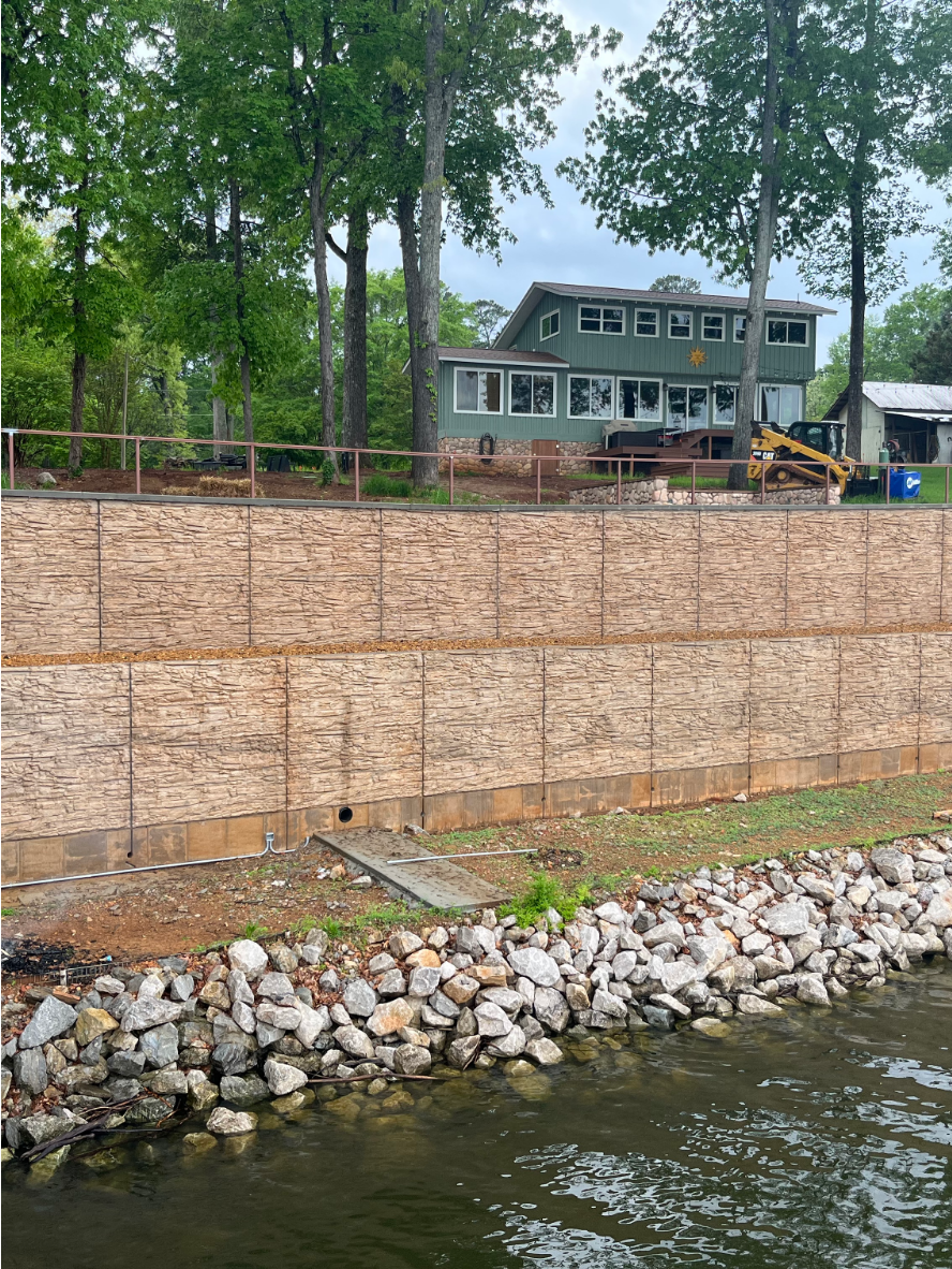 large retaining wall finished with Caf Royale EasyTint Large lakeside concrete retaining wall stained with Café Royale EasyTint™, featuring warm tan and brown tones that mimic natural stone under a tree-lined landscape.