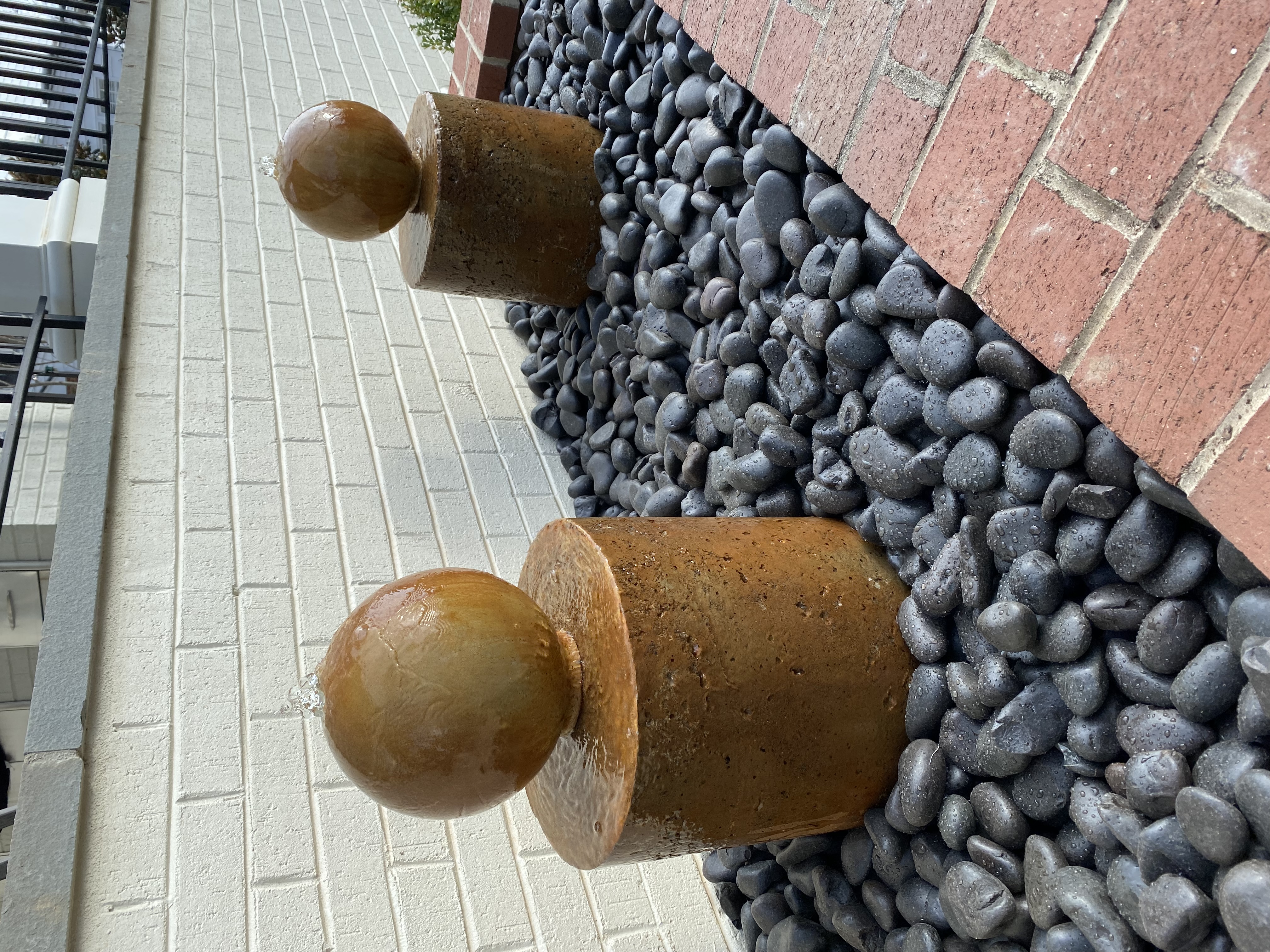 A photograph of the completed DIY concrete fountain, featuring a stained spherical piece atop a cylindrical stand, both richly colored with Malayan Buff and Desert Amber acid stain, set on a bed of gray river rocks.